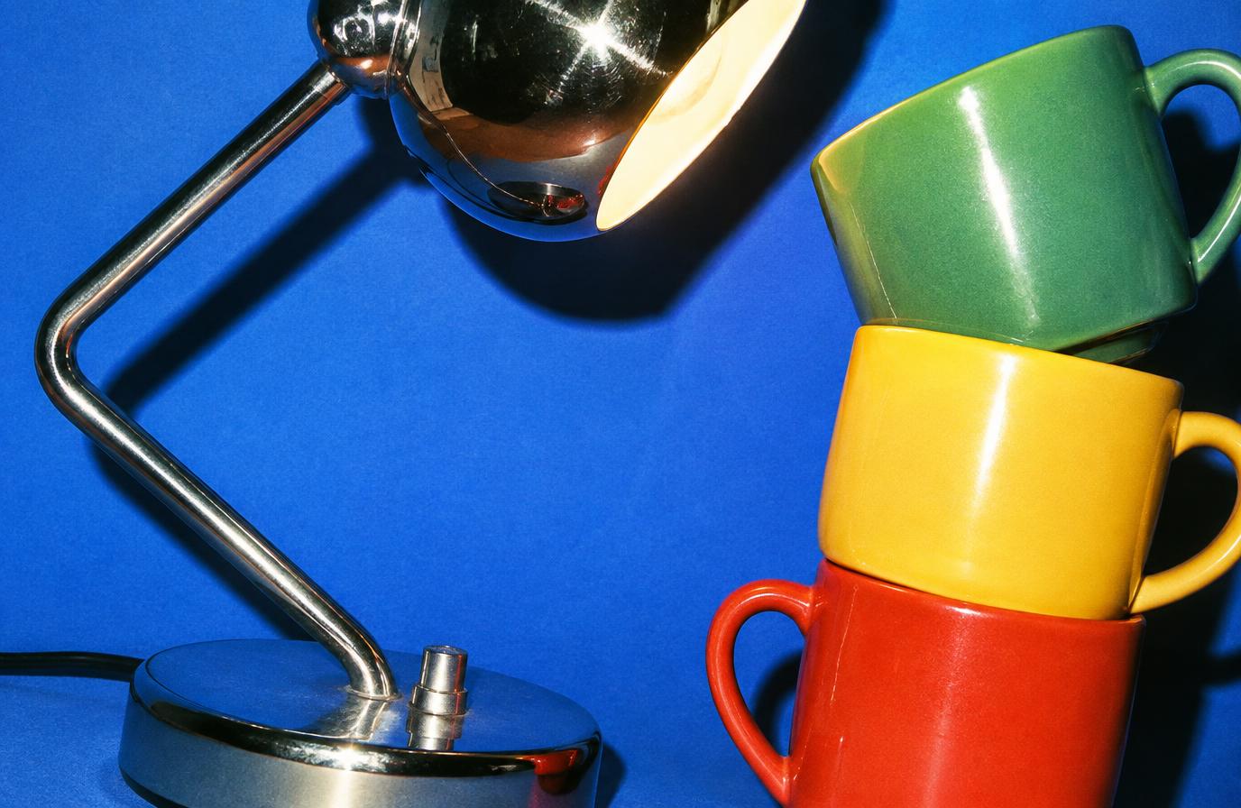 Chrome desk lamp and a leaning stack of red, yellow and green mugs on a cobalt blue background